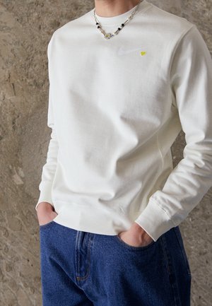 Hombre vistiendo sudadera blanca con pequeño logo de corazón amarillo y collar de cuentas, manos en los bolsillos de jeans azules contra fondo de pared de piedra.