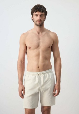 Hombre sin camiseta con cabello corto y oscuro que lleva pantalones cortos crema con textura y cordón, de pie frente a un fondo claro y liso, mirando al frente.