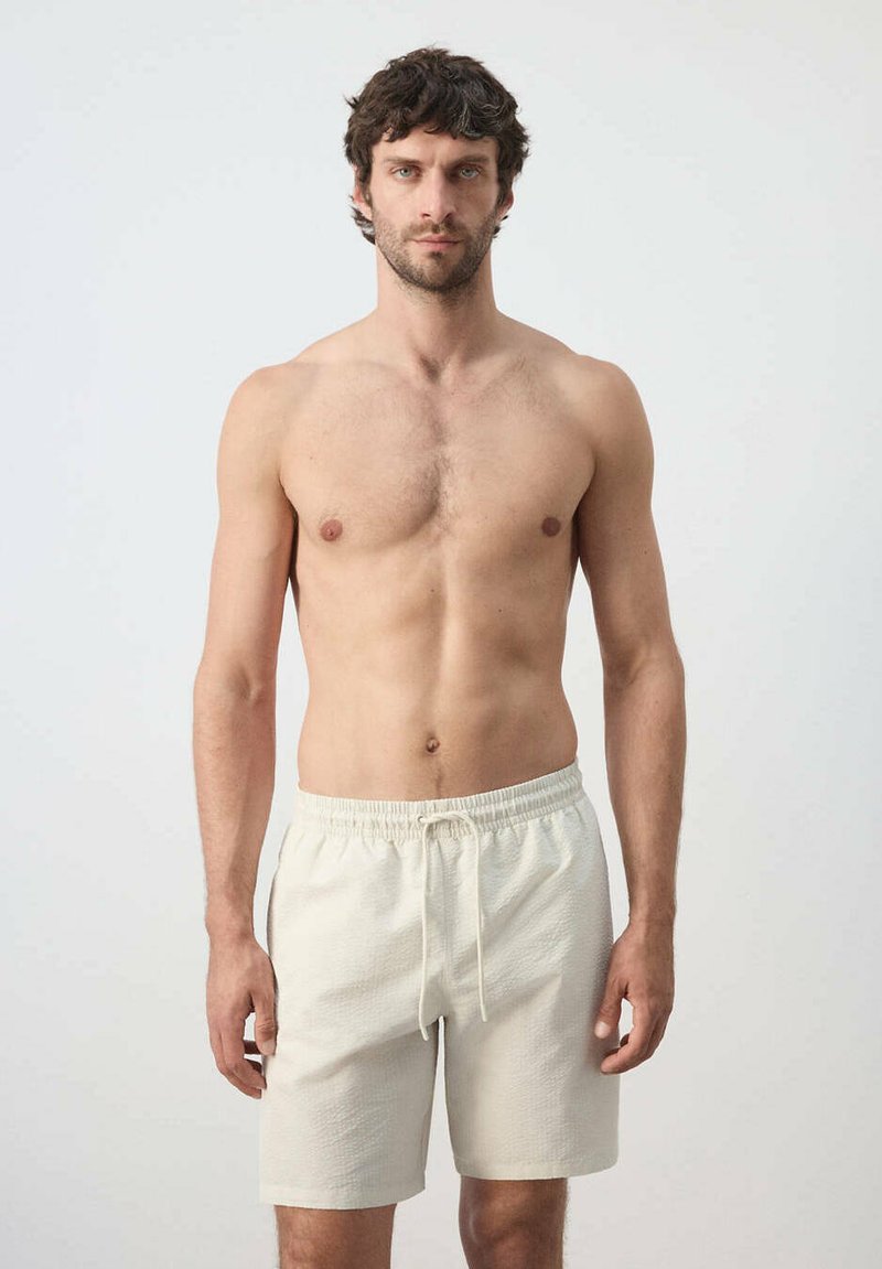 Homme torse nu aux cheveux courts et foncés portant un short crème texturé avec un cordon, debout devant un fond clair uni, faisant face devant.