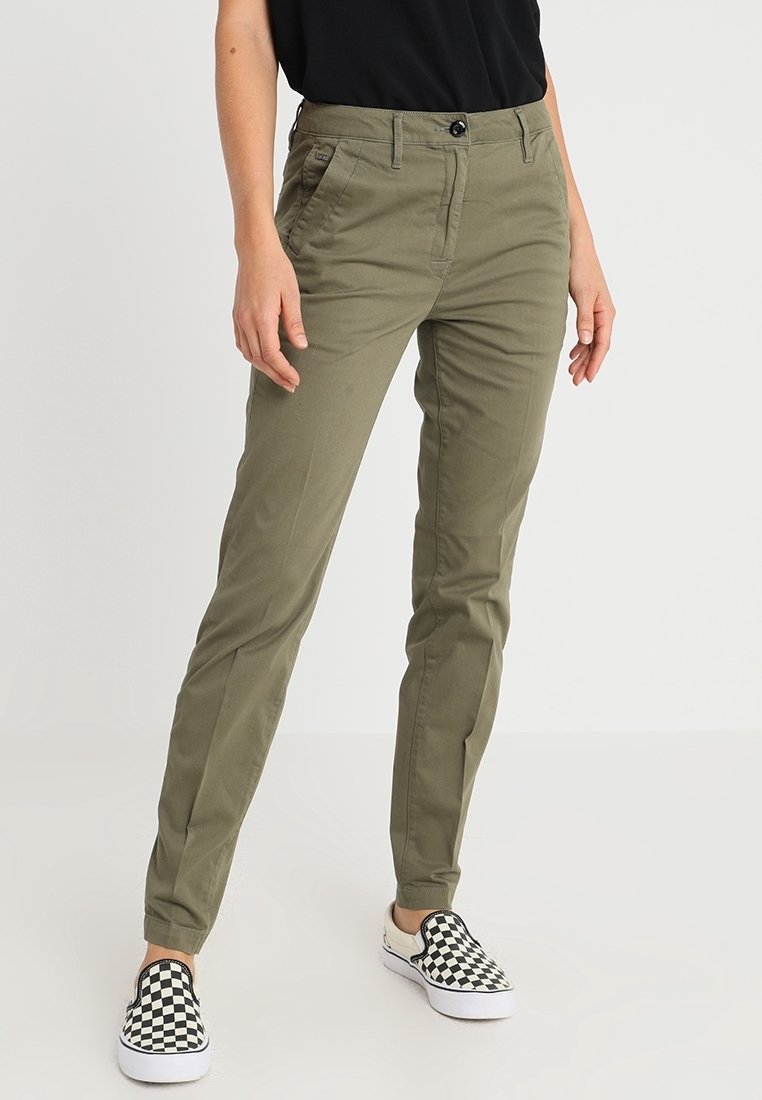 Personne portant un pantalon slim kaki, un haut noir et des chaussures slip-on à carreaux noir et blanc, debout devant un fond clair uni.