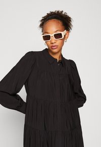 Svart långärmad klänning med knäppning framtill, plisserade sektioner och böjda sömmar. Fyrkantiga, ljusfärgade solglasögon. Slät textur.