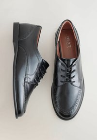 Une paire de chaussures de ville pour hommes en cuir noir poli, avec des lacets et des détails en brogue, sur une surface claire.