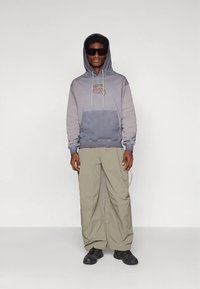 Ανδρας στέκεται φορώντας γκρι φούτερ με κουκούλα και λογότυπο, χαλαρούς μπεζ παντελόνες, μαύρα παπούτσια και μεγάλα μαύρα γυαλιά ηλίου, μπροστά σε ουδέτερο φόντο.