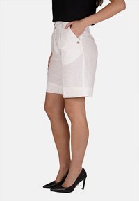 Pantaloni corti bianchi in lino con una vestibilità rilassata, pieghe, tasche laterali e chiusura con zip. Indossati con tacchi neri a punta, che mettono in risalto una silhouette elegante.