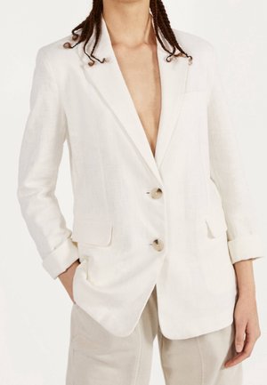 Lněný blazer krémové barvy s hlubokým výstřihem do V, dvěma předními kapsami a velkými knoflíky. Má ohrnuté rukávy a volný střih.