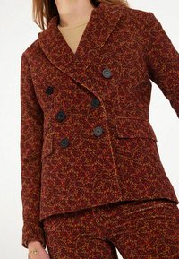Blazer double boutonnage en tissu texturé rouge et or à motifs paisley, avec boutons noirs et poches à rabat à l'avant. Design de col élégant.