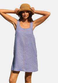 Helllila Leinenkleid mit quadratischem Ausschnitt, das eine zentrale Naht und keine Ärmel aufweist. Kombiniert mit einem strukturierten Strohhut.
