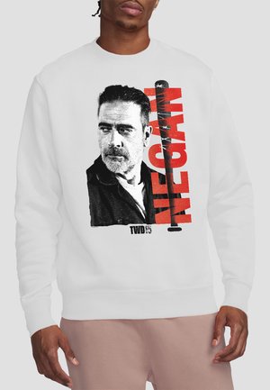 Mann trägt weißen Pullover mit schwarz-weißem Porträt eines bärtigen Mannes und großem rotem Text "NEGAN" mit Stacheldraht-Design.