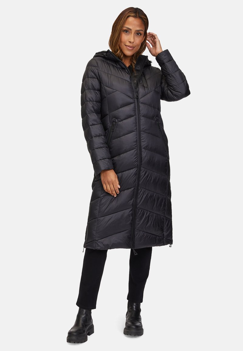 Lange zwarte pufferjas met een capuchon, voorzien van een rits aan de voorkant, een chevronpatroon en twee zijzakken met rits. Draagt over zwarte broek en laarzen.