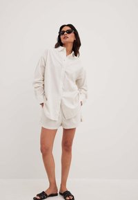 Lightweight, striped button-up shirt in white and beige, paired with matching shorts. Worn with black sunglasses and sandals.