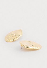 Zlaté kovové náušnice s vlnovitým dizajnom a lesklým povrchom a jemnými textúrovými detailmi. Každá náušnica má nepravidelný tvar.