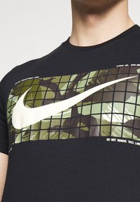Camiseta negra de manga corta con un gran gráfico de patrones en verde y negro, con el logotipo de Nike en blanco centrado en el pecho.