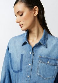 Camicia in denim di colore azzurro chiaro con colletto a punta, bottoni a snap e tasca sul petto. Presenta dettagli di cucitura distintivi sul davanti.
