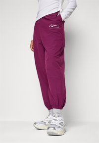 Person i lila Nike Sportswear joggers, vit långärmad tröja, grå strumpor och vita och silverfärgade Nike-skor, står med händerna i fickorna.