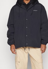 Man som bär en svart Carhartt-hoodie med tryckknappar och beiga cargobyxor, stående mot en enkel bakgrund.
