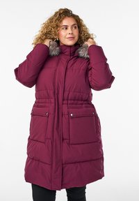Sieviete valkā garu purpura puffer mēteli ar mākslīgās pūkas apdari, pielāgojot apkakli, nedaudz skatoties sānis, stāvot pret baltu fona.