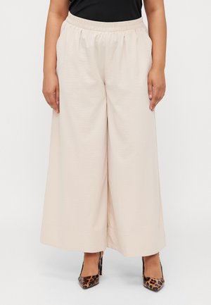 Pantalons larges beige avec une taille élastique et des poches ; fabriqués en tissu léger et texturé. Associés à des talons imprimés léopard.