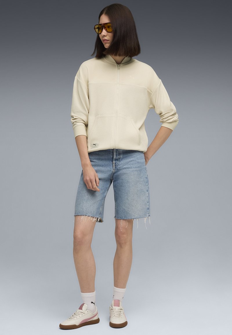 Kvinde iført cremefarvet zip-up jakke, lyseblå denimshorts, hvide sokker, cremefarvede sneakers og solbriller med orange toning mod en grå baggrund.