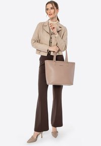 Beige totebag met een gladde textuur en gouden accenten, gehouden in de hand van een vrouw. Ze draagt een beige jas en een brown broek.