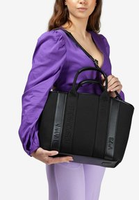 Bolso tote negro con texto en relieve y asas negras, con una superficie texturizada. Llevado en el brazo, junto a una blusa morada.