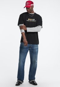 T-Shirt nera con maniche grigie, caratterizzata da un logo "GUESS JEANS COMPANY" in grafica multicolore. Indossata con jeans blu strappati e scarpe nere.