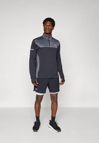 Conjunto desportivo masculino composto por uma camisola de manga comprida cinzento escuro com gola com fecho éclair e detalhes em cinzento claro, combinado com calções pretos e sapatilhas pretas.