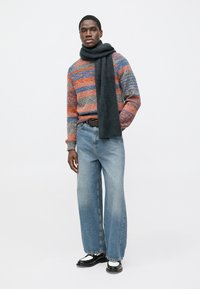Farebný pruhovaný sveter v oranžovej, sivej a modrej, kombinovaný so svetlomodrými džínsami s širokými nohavicami. Okolo krku je prehodená tmavá šatka. Čierne topánky.