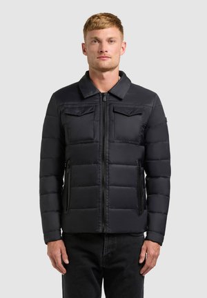 Bărbat blond purtând o geacă neagră matlasată cu fermoar frontal, buzunare la piept cu pată și buzunare laterale cu fermoar, stând în fața unui fundal simplu.