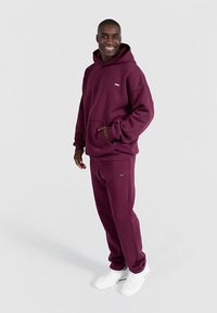Bordowa bluza z kapturem i dopasowane spodnie dresowe z miękkiej, teksturowanej tkaniny. Z kieszenią typu kangur, małym logo i w połączeniu z białymi trampkami.
