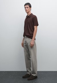 Hnědé polo tričko, světle šedé džíny a tmavé mokasíny. Model stojí na jednobarevném pozadí, předvádějící jednoduchý casual outfit.