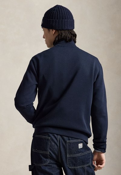 Man wearing a navy blue knit beanie, navy zip-up sweater, and dark denim pants, shown from the back against a neutral background.