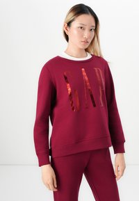 Burgundy sweatshirt with shiny "GAP" logo in metallic rose gold. Features crew neck, ribbed cuffs, and hem; made of textured fabric.