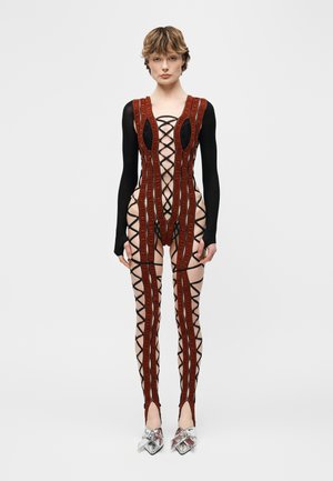 Nő fekete és piros fűzős bodyban, áttetsző panelokkal és hosszú ujjakkal, ezüst hegyes orrú cipővel párosítva.