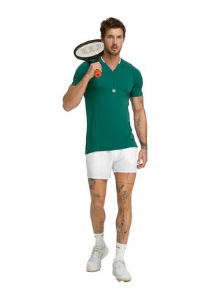 Polo vert ajusté à manches courtes avec des accents blancs, associé à un short blanc. Le modèle tient une raquette de tennis.