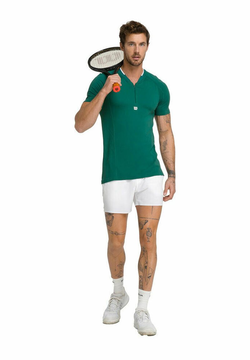 Polo shirt verde aderente con maniche corte e dettagli bianchi, abbinato a pantaloni corti bianchi. Il modello tiene in mano una racchetta da tennis.