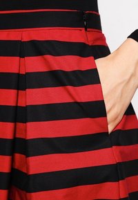Les mains reposent dans la poche d'une jupe rayée horizontale rouge et noire, avec une fermeture éclair visible et des plis près de la taille.