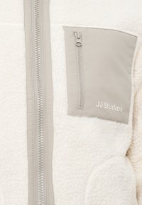Veste en polaire crème avec une poche poitrine zippée grise. Présente une fermeture éclair lisse à l'avant et un tissu doux et texturé. Marquage JJ Studios visible.