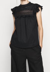 Mujer vistiendo una blusa sin mangas negra con detalle de encaje en la parte superior y hombros con volantes, combinada con pantalones negros, con las manos en los bolsillos.