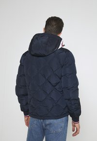 Man som bär en marinblå kviltad puffjacka med huva över blå jeans, stående med ryggen mot kameran mot en enkel vägg.