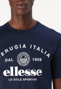 Tamsiai mėlynas medvilninis marškinėliai su apvaliu kaklu, su baltais užrašais ir logotipu „ellesse“, ir „Perugia Italia DAL 1959“ drąsiais rašmenimis.
