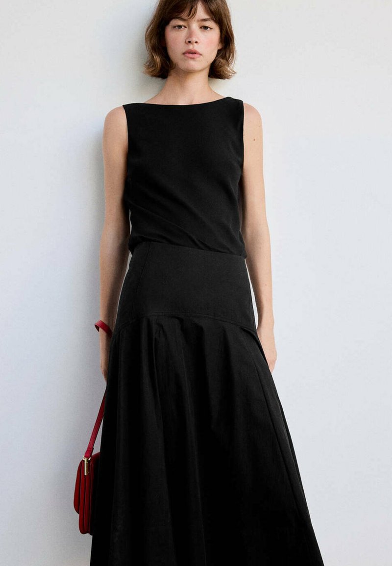 Jeune femme portant un haut noir sans manches et une jupe noire taille haute, tenant un sac à main rouge, debout contre un mur clair uni.