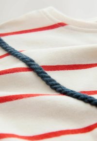 Gestreifter Baumwollstoff mit roten Linien auf cremefarbenem Hintergrund, akzentuiert durch eine gedrehte blaue Seilverzierung entlang der Naht.