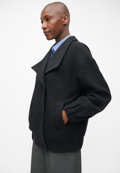 Chaqueta de doble botonadura de mezcla de lana negra con grandes solapas, bolsillos laterales, puños acanalados y un acabado texturizado. Lleva puesta sobre una camisa a rayas azul y blanca.