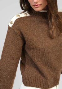 Pull marron tricoté avec un col montant, un détail d'épaule blanc contrastant et des boutons décoratifs dorés sur les épaules.