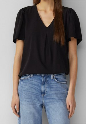 Kvinde iført en sort V-hals bluse med korte puffærmer og lyseblå høj talje jeans, stående mod en ensfarvet baggrund.
