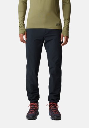 Mann trägt schwarze Outdoorhose, schwarze Wanderschuhe mit roten Schnürsenkeln und ein olivgrünes Langarmshirt mit Daumenlöchern.