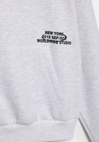 Sivá mikina s jemnou textúrou. Obsahuje predné vrecko a čiernym vyšitým textom: "NEW YORK 2018 SEP/O3 WORLDWIDE STUDIO."