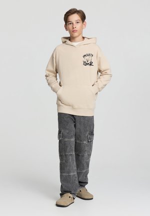 Felpa beige in cotone con tasca frontale e stampa grafica nera. Pantaloni cargo grigi con più tasche. Scarpe slip-on beige.