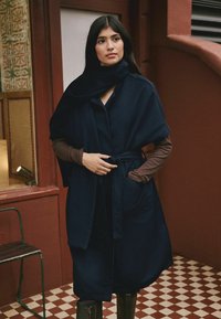 Woman with long dark hair wears a navy belted wool cape and brown long-sleeve top, standing on checkered floor beside red and patterned walls.