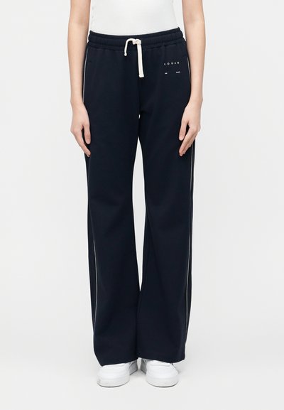 Ενήλικας που φοράει μαύρο φαρδύ παντελόνι φόρμας με λευκό κορδόνι, μικρό λευκό λογότυπο κοντά στη μέση, σε συνδυασμό με λευκά αθλητικά παπούτσια και λευκή μπλούζα.